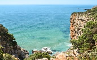 Dramatiske klippeskrænter dækket af grøn vegetation med udsigt over turkisblåt vand langs Algarvekysten mellem Lagos og Tavira, Portugal.