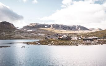DNT Finsehytta hyttekompleks på Hardangervidda-plateauet ved en bjergsø med snedækkede toppe i baggrunden under skyet himmel.