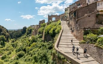 Group of hikers walking on curved stone path leading up to historic Orvieto town with ancient buildings and green valley below under blue sky.