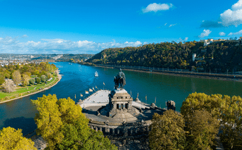 Luftfoto af Deutsches Eck monument i Koblenz, hvor Rhinen og Mosel mødes, med rytterstatue, grønne træer og fæstningsbakke.