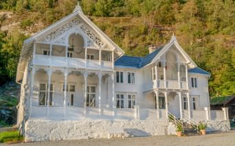 Hvidt to-etagers træhotel med udsmykkede detaljer og balkoner ved en grøn skråning i Norge.