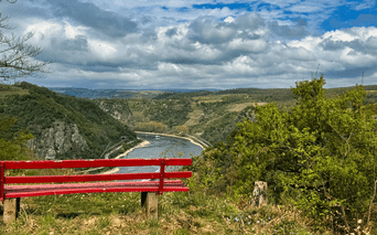 Rød træbænk på Rheinsteig med udsigt over Rhinen, der slynger sig gennem grøn dal med bakker under overskyet himmel.