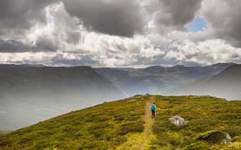 Vandrer i blå jakke på græsklædt bjergsti langs Kongevegen i Norge, med dramatisk overskyet himmel og tåget dal.