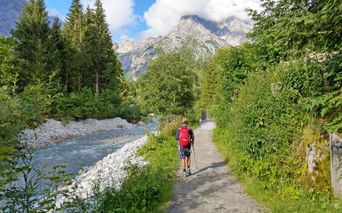 Hiker with red backpack walking on gravel path beside turquoise river in Austrian Alps, surrounded by green forest and rocky peaks.