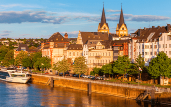 Aftenvisning af Koblenz flodfront med historiske bygninger og kirke med tvillingetårne. Et krydstogtskib er fortøjet langs promenaden.
