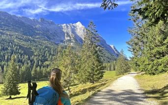 Vandrer med blå rygsæk på grusvej gennem alpeng. Bjergtinder og nåleskov synlige i baggrunden under blå himmel.