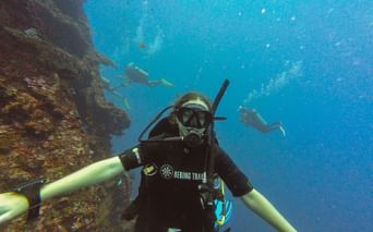 Undervandsselfie af dykker i sort våddragt med Bering Travel-logo, der udforsker koralrev i Balis klare blå vand med andre dykkere synlige.