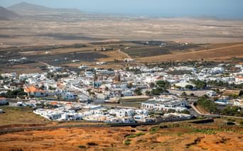 Aerial view of Teguise, a white-walled town on Lanzarote island, surrounded by volcanic landscape with mountains in the background.