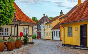 Kullerstensgata i Odense med färgglada korsvirkeshus, gula och vita fasader och röda tegeltak under blå himmel.