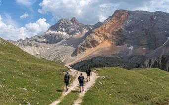 Tre fotturister med sekker går på en jordsti gjennom grønne alpine enger i Dolomittene, med dramatiske kalksteinstopper i bakgrunnen.