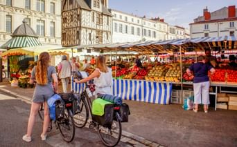 Udendørs marked i La Rochelle med frugt- og grøntsagsboder. To cyklister med cykeltasker ser på markedet foran historiske bygninger.