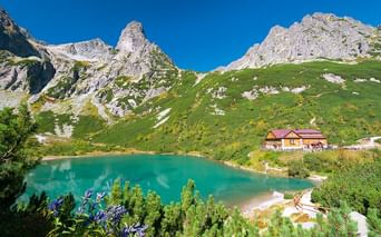 Turkis bjergsø i Tatrabjergene med en gul hytte med rødt tag ved bredden, omgivet af grønne skråninger og klippefyldte toppe.