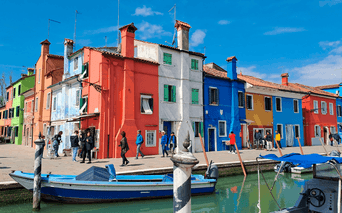Række af farverige huse i rødt, hvidt, grønt, blåt og orange langs en kanal med fortøjede både i Venedig under blå himmel.
