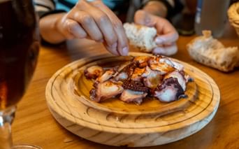 Grillede blekkspruttentakler servert på en rund tretallerken. Hender som holder brød er synlige i bakgrunnen på et trebord.