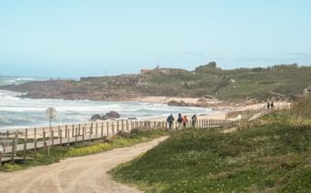 Pilgrims walking on sandy path with wooden fence along Atlantic coast on Portuguese Camino. Green vegetation and beach visible with ocean waves.