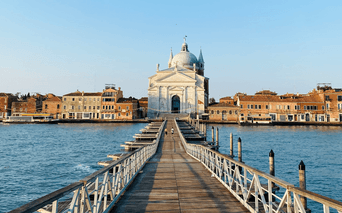 Lang træbro over blåt vand til hvid kuppelkirke i Venedig. Historiske bygninger langs vandkanten under klar himmel.