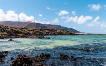 Volcanic coastline near Orzola, Lanzarote with turquoise water, black lava rocks, and barren hills under a blue sky with white clouds.