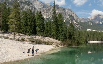 Vandrere ved Dolomitternes bjergsø Tre vandrere på en stenstrand ved en turkis bjergsø i Dolomitterne, med dramatiske kalkstenstinder og nåleskov.
