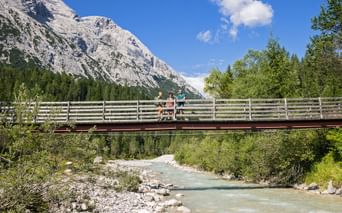 To vandrere på en træbro over en turkis bjergbæk, med klippefyldt Zugspitze-top og grøn skov i baggrunden.
