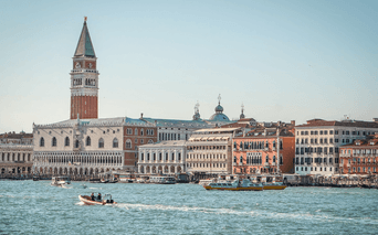 Venedigs vandkant med Markus Kampanile klokketårn, historiske bygninger og både på vandet. Klar himmel over den venetianske lagune.