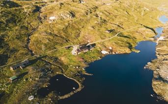 Luftfoto af Geiterygghytta DNT-hytte på Hardangervidda, beliggende på en halvø mellem blå bjergsøer omgivet af gylden tundra.
