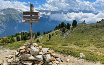 Träskylt på stenkummel i alpin äng med bergstoppar och moln i bakgrunden på vandringsleden Garmisch till Merano.