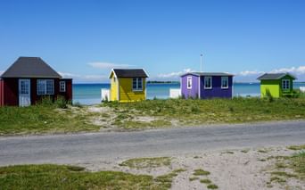 Fyra färgglada strandstugor i rött, gult, lila och grönt längs en sandstrand på ön Ærø med blått hav och klar himmel i bakgrunden.