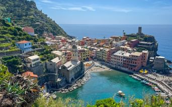Aerial view of Vernazza, a colorful coastal village in Cinque Terre with a small harbor, historic tower, and buildings nestled between hillside and sea.