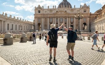 Two hikers with backpacks raising arms in celebration on St. Peter's Square in Rome, with St. Peter's Basilica and colonnade in background.