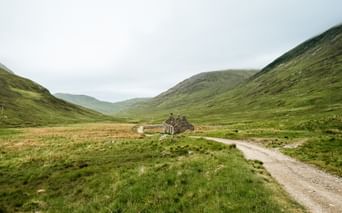 Grusvej der fører til stenruin i grøn dal på West Highland Way, Skotland. Bølgende bakker indramme dalen under overskyet himmel.
