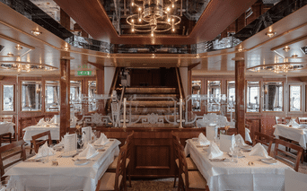 Elegant skibsrestaurant med hvide duge, træpaneler og central trappe. Reflekterende loft og panoramavinduer langs væggene.