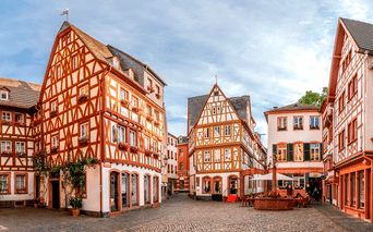 Brostensplads i Mainz omgivet af farverige bindingsværkshuse med røde og hvide facader. Udendørs siddepladser synlige på pladsen.