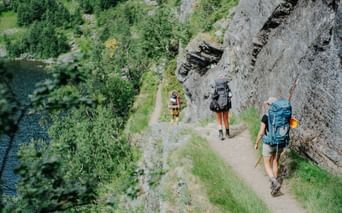 Tre vandrere med rygsække på en smal bjergsti i Aurlandsdalen, Norge. Klippevæg til højre, grøn dal med flod nedenfor.