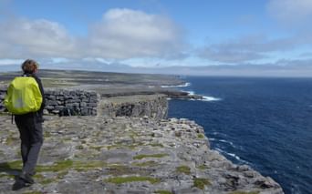 Person i lysegul jakke står på kalkstensklipper med udsigt over Atlanten. Stenmure og klippefyldt terræn synligt ved Irlands kyst.