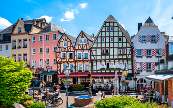 Farverige bindingsværkshuse og traditionelle bygninger omkring en offentlig plads i Linz med springvand, udendørs siddepladser og blå himmel.