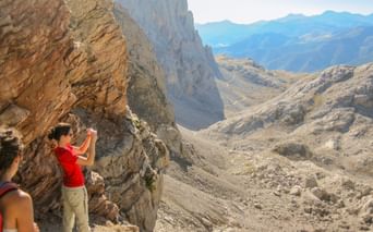To vandrere på en klippefyldt bjergsti i Picos de Europa, Spanien. En person i rødt fotograferer de dramatiske kalkstenstinder.
