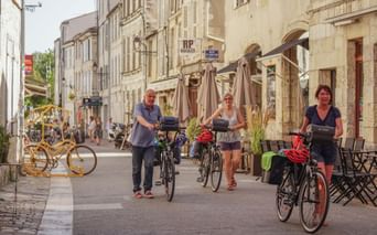 Tre cyklister med cykler går gennem en gågade i historiske La Rochelle, Frankrig. Stenbygninger med caféer langs begge sider.