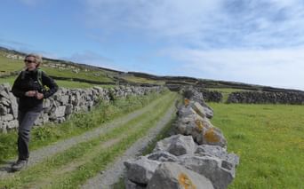 Vandrer med solbriller på jordvej ved stenmure i irsk landskab. Grønne marker og bakkede landskab under skyet himmel.