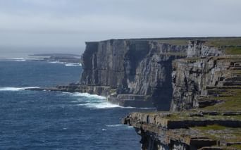 Høje kalkstensklipper langs Irlands vestkyst med lagdelte klippeformationer. Hvide bølger bryder mod de mørke klipper.
