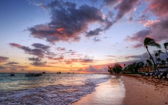 Dramatisk solnedgang over en strand på Bali med palmer i vinden, fargerike skyer reflektert på våt sand og båter utenfor kysten.