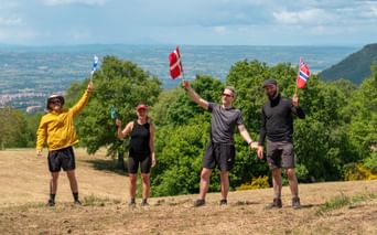 Four hikers holding flags from different countries on a hilltop along Via Francigena near Orvieto, with green hills and valleys in background.