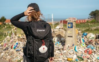 Rear view of pilgrim with backpack and scallop shell on Portuguese Camino, walking past stone markers with yellow arrows toward coastal village.