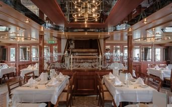 Elegant skibsrestaurant med hvide duge, træstole og historisk skovlhjul som midtpunkt. Spejlloft og varm belysning.
