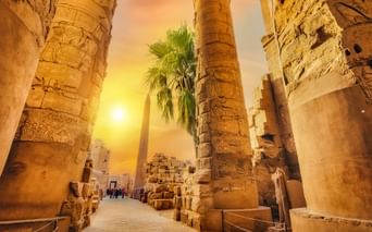 Massive stensøjler med hieroglyfiske udskæringer ved Karnak-templet i Egypten, oplyst af gyldent solnedgangslys med palmer.