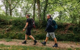 To vandrere med rygsække går på en skovsti kantet af grønne træer på Caminoen fra Sarria til Santiago.