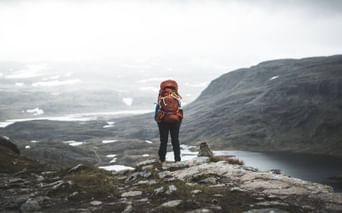 Vandrer med orange rygsæk på klippeformation i Skarvheimen, Norge, med udsigt over bjergplateau med søer og snefelter.