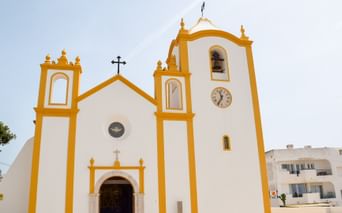 Hvid portugisisk kirke med gule detaljer, to klokketårne og et ur. Kirken har et kors på toppen i Burgau, Lagos.