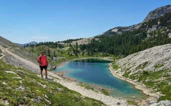 Vandrare i röd tröja ser ut över turkos alpin sjö i Juliska Alperna, Slovenien. Klippiga sluttningar och skogsklädda berg omger vattnet.