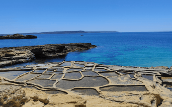 Traditionelle saltpander udskåret i kystklipperne på Gozo, Malta, med turkisblåt Middelhav og fjerne klipper under klar blå himmel.