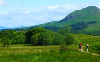 Fotturister på West Highland Way Skottland To fotturister går på en sti gjennom de grønne skotske høylandet med bølgende åser og fjell i bakgrunnen under en skyet himmel.